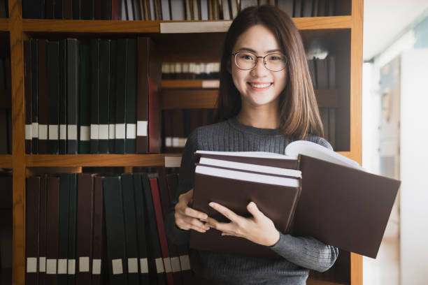 educação primeiro, beautiful feminino estudante universitário segurando livros dela sorrindo alegremente em pé na biblioteca, educação aprendizagem alta escola programa adolescente inteligente conceito, conceito de aprendizagem e educação - registo livro - fotografias e filmes do acervo