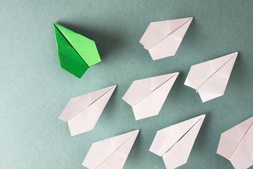 Green paper plane is flying to the left on blue background where a group of white paper planes are flying to the right.