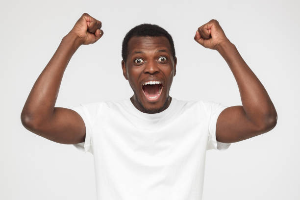 istock Young african american man in blank white t shirt, shouting while his team win, raised both fist in victory gesture, isolated on gray background 1000221524