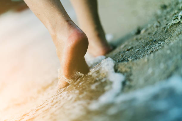 istock closed up barefeet walk alone on beach sand ocean water with foam 1000406572
