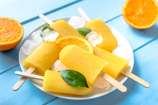 istock Orange popsicle on blue wooden background. Summer refreshing ices on stick. 1000595506
