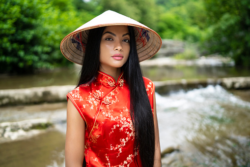 Elegant young woman in traditional chinese dress standing on river, looking at camera.