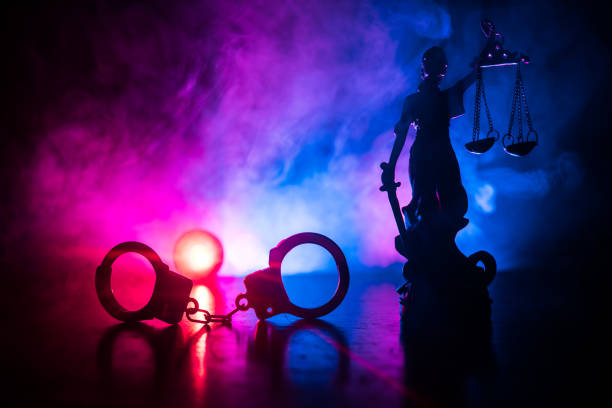 Legal law concept. Silhouette of handcuffs with The Statue of Justice on backside with the flashing red and blue police lights at foggy background. stock photo