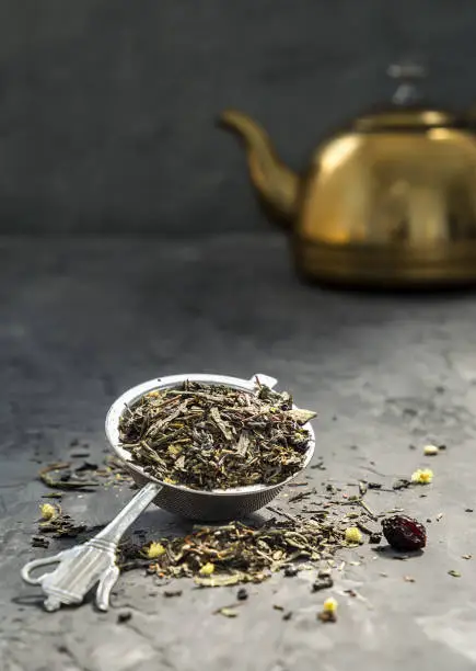 Herbal tea in metal spoon against dakr background of a yellow teapot, Dark style. Copy space