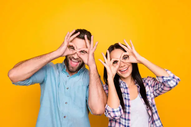 Photo of Joke humor ha-ha concept. Portrait of funky cheerful couple making binoculars with fingers wearing casual shirts isolated on bright yellow background