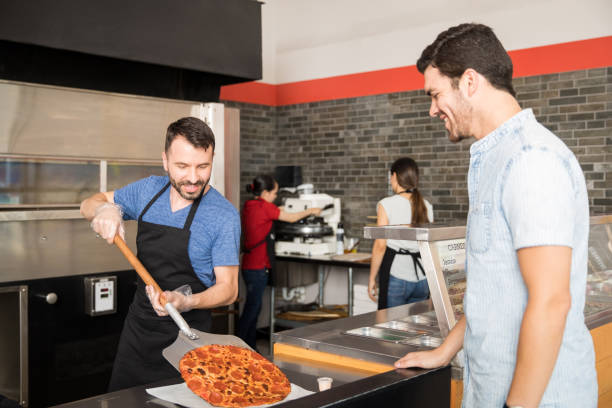 souriant chef mettre la pizza sur la plaque à l’aide de pelle, alors que le client regarde - pizza pizzeria restaurant waiter photos et images de collection
