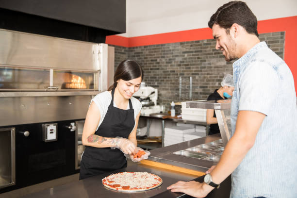 istock Smiling woman adding pepperoni slices to cheese pizza in kitchen counter 1000842712