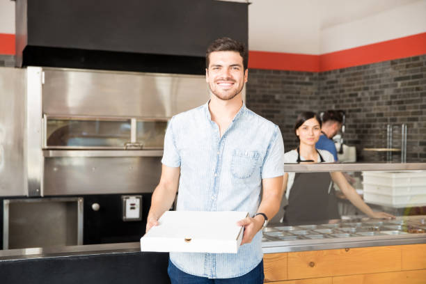 souriant jeune homme tenant la boîte à pizza au comptoir de la pizza - pizza pizzeria restaurant waiter photos et images de collection