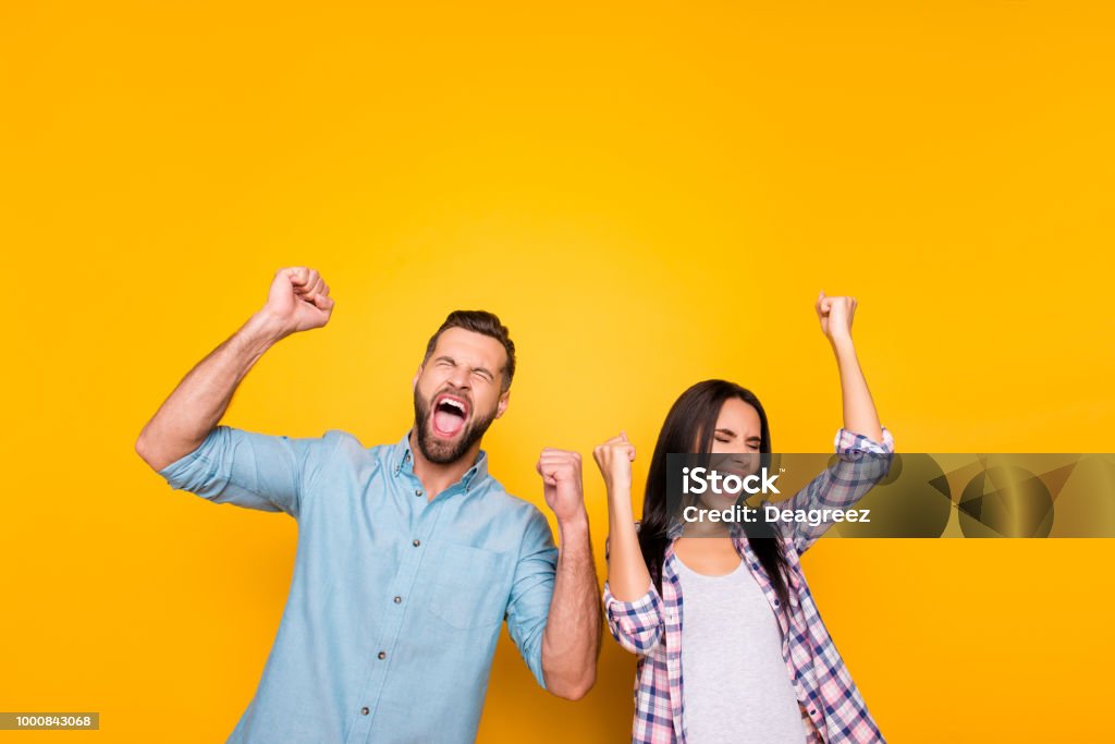 Portrait of crazy man couple full of happiness yelling loudly holding raised arms keeping eyes closed celebrating victory isolated on vivid yellow background Celebration Stock Photo