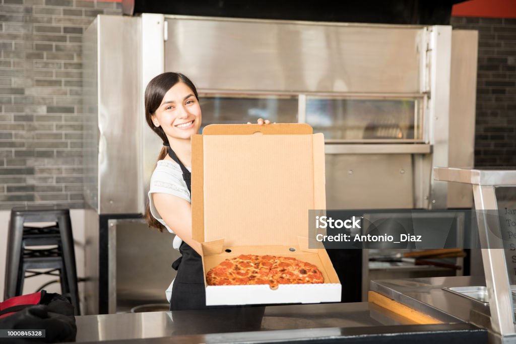 Young smiling chef with pizza in open box Latin woman chef wearing black apron holding freshly baked pizza in open box at pizza shop counter looking at camera Pizzeria Stock Photo