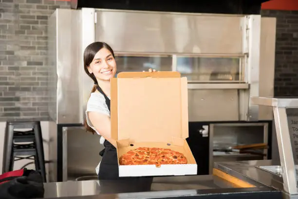 Latin woman chef wearing black apron holding freshly baked pizza in open box at pizza shop counter looking at camera