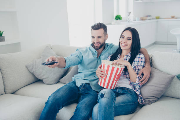 portrait of cheerful joyful partners embracing sitting on sofa searching interesting program with remote control ready for watching comedy having snack bucket of popcorn - remote television movie box imagens e fotografias de stock