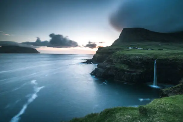 A waterfall under a little village flowing down green cliffs into the blue deep sea with a dramatic sky and clouds during sunset on the Faroe Islands
