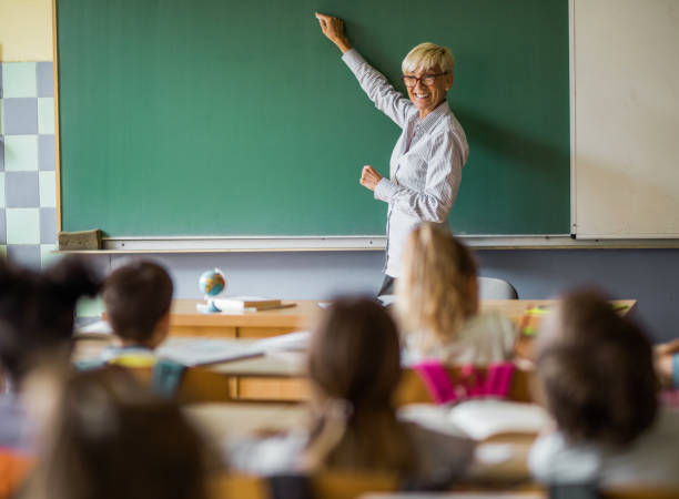 Happy senior teacher writing a lecture on blackboard at elementary school. Happy senior teacher teaching elementary students on a class in the classroom and writing on blackboard. Copy space. elementary school building photos stock pictures, royalty-free photos & images