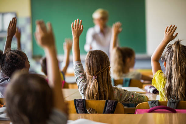 Back view of elementary students raising their arms on a class. Rear view of large group of students raising their arms to answer the question on a class at elementary school. elementary school stock pictures, royalty-free photos & images