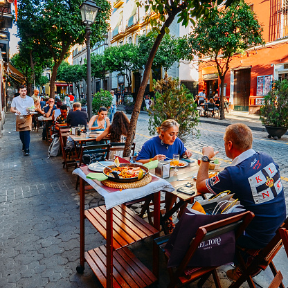 Sevilla, Spain - July 15, 2018: Tourists enjoy at restaurant terrace typically Spanish cuisine, seafood paella with squids, mussels, clams, and prawns on a bed a saffron flavoured rice