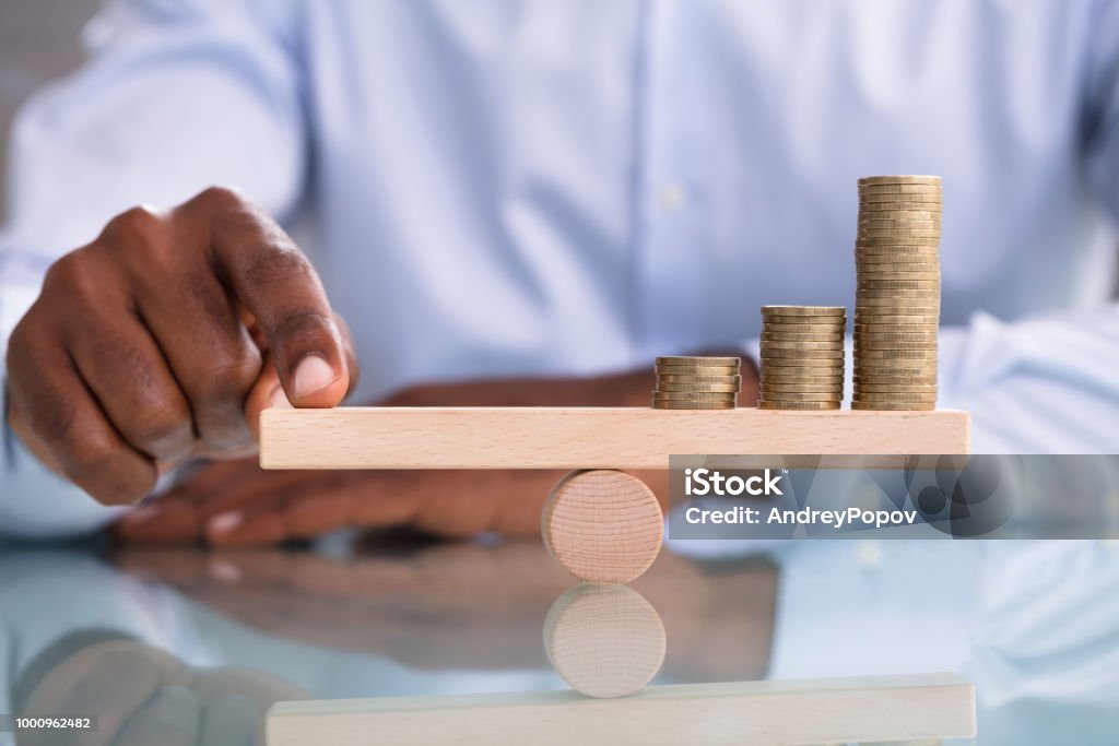 Businessman Balancing The Coin Stack On Wooden Seesaw Close-up Of A Businessman Balancing The Coin Stack On Wooden Seesaw Over The Reflective Desk Currency Stock Photo
