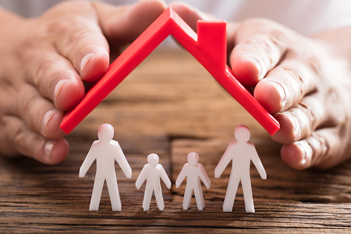 A person's hand protecting family figures with red roof on wooden desk