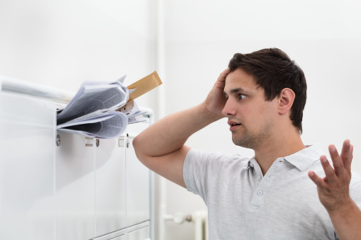 Close-up of man with hand on head in front of overloaded mailbox with junk mail