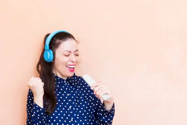 Photo of beautiful arab girl wearing blue t-shirt  she singing, holding a remote control tv and listening to music with  wireless earphones isolated over orange wall