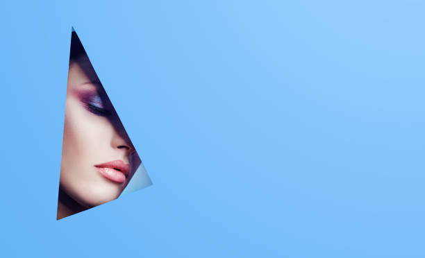 istock Woman with a beautiful look through a triangular hole in the blue paper, bright makeup and pink lipstick. Advertising cosmetics, professional art makeup, lip gloss, bright color, blue background 1001075690