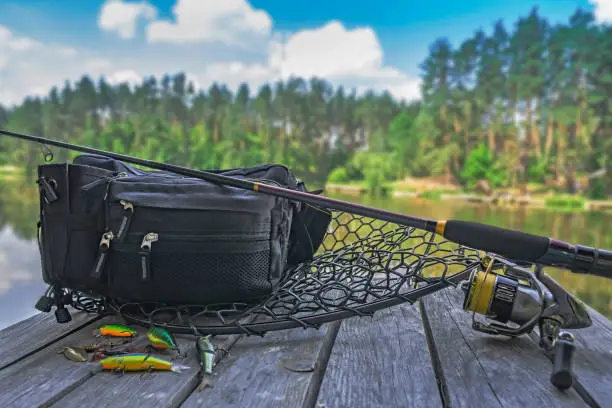 Photo of Fishing tackle set. Spinning rod with reel and lures on wooden platform on forest lake background
