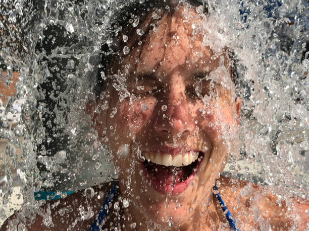 A woman, in summer, stands underneath a fountain and is splashed by cool water.