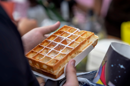 Belgian fresh warm waffle with powdered sugar in the hands of the buyer. Gastronomic dainty products on market counter, real scene in food market