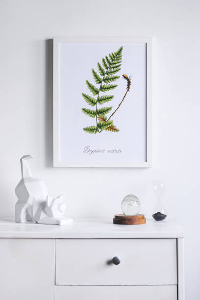 istock Stylish and modern white interior with cupboard, cat figures, mock up poster frame and hourglass. Scandinavian white home interior. 1001156684