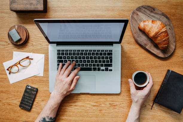 Modern and stylish composition of wooden desk with laptop, croissant, cup of coffee, phone, notebook, glasses and office accessories. Hands writing on laptop keyborad. Creative desk .Flat lay. Concept of stylish flat lay desk. keyborad stock pictures, royalty-free photos & images