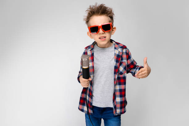 A handsome boy in a plaid shirt, gray shirt and jeans stands on a gray background. A boy wearing sunglasses. The boy holds a microphone in his hand. The boy is reading rap Charming happy child on gray background. The boy's hair is up. The boy has a hairstyle. The boy in round sunglasses. A handsome boy with a red hair color. The boy holds a microphone in his hand. rap kid stock pictures, royalty-free photos & images