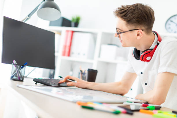 istock A young man in glasses stands near a computer desk. A young man works with a computer and a magnetic board. On the neck, the guy's headphones hang. 1001165232