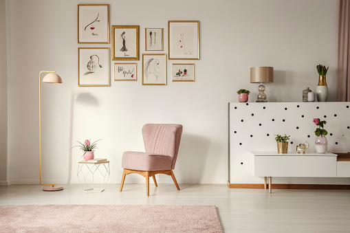 Old-fashioned armchair, pastel pink floor lamp and stylish, golden decorations in a retro living room interior with white walls