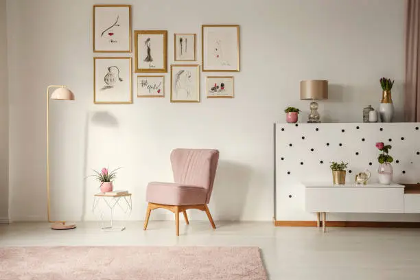 Photo of Old-fashioned armchair, pastel pink floor lamp and stylish, golden decorations in a retro living room interior with white walls