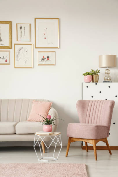 istock Book and plant on a diamond shape side table, beige sofa and comfy armchair in an elegant, white living room interior with framed posters gallery 1001171316