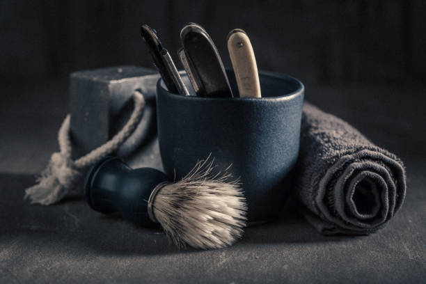 A young and handsome man with a beard is preparing shaving cream with a brush in a bowl. In front of a wooden wall.