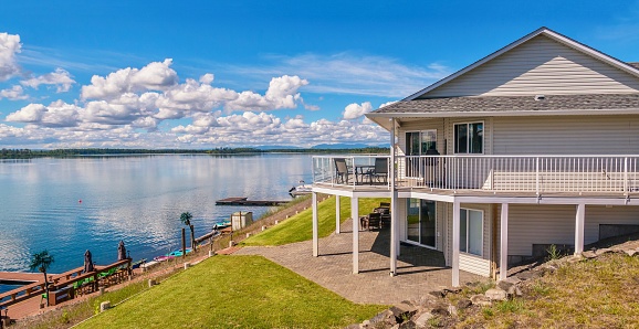 A large two-story house with beige vinyl siding, a shingled roof, a large outdoor deck and patio, with outdoor furniture situated on a freshwater lake. There is a a dock with a sitting area and equipment for water sports.