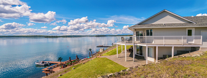 A large two-story house with beige vinyl siding, a shingled roof, a large outdoor deck and patio, with outdoor furniture situated on a freshwater lake. There is a a dock with a sitting area and equipment for water sports.