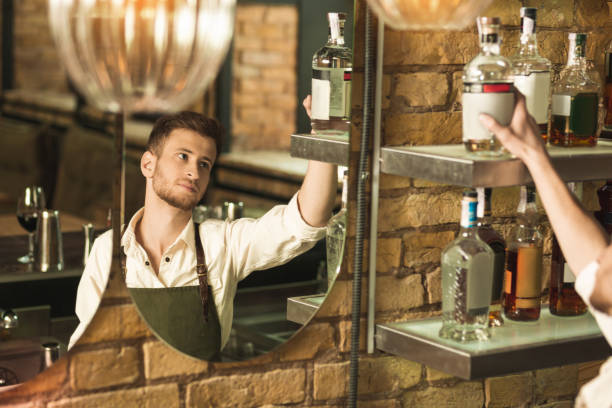 Handsome young barman taking bottle of alcohol Diligent worker. Handsome young barman taking a bottle of alcohol from the shelf while mixing a cocktail restaurant mirror stock pictures, royalty-free photos & images