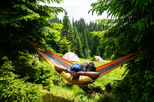 Adventurer relaxes in hammock on the green mountain meadow among fir trees and admires view. Epic travel in the mountains. Back view.