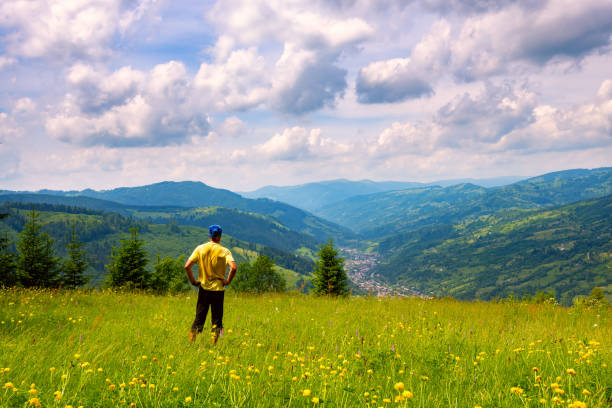 Adventurer is standing on the green mountain meadow among flowering dandelions and admiring mountain valley stretching to the horizon in front of him. Back view.