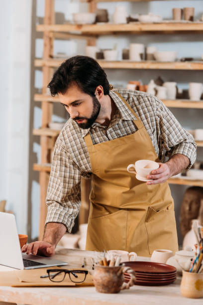 istock male potter in apron holding ceramic cup and using laptop in workshop 1001342748