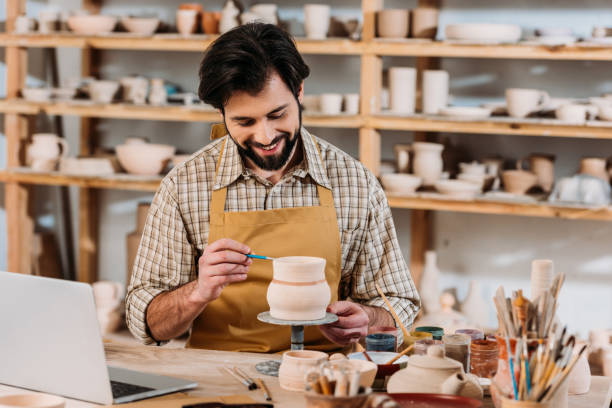 istock smiling potter painting ceramic jug in workshop with laptop 1001349270