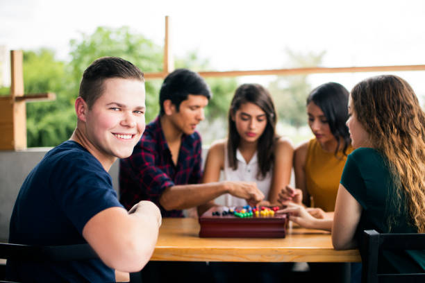 A young caucasian man sitting with friends at a table, playing with a board game and smiling at the camera.
