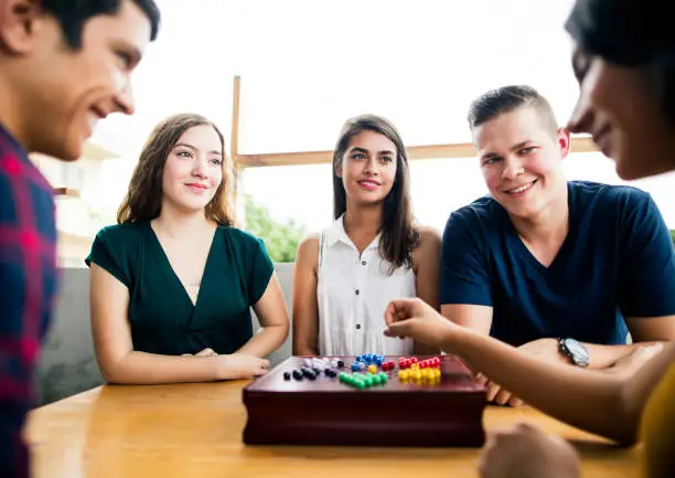 A small group of cheerful young people sitting at a table, playing with a board game and smiling at each other.