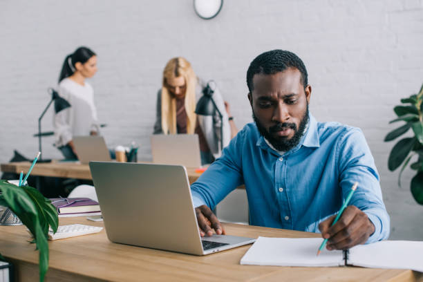 istock young african american businessman writing in textbook and using laptop at table and two businesswomen working behind 1001354426