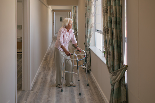 Shot of a senior woman using a walker and looking thoughtfully out of a window in a retirement home
