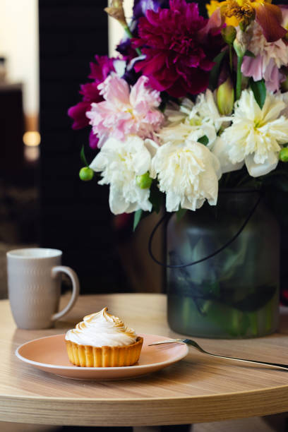 Lemon curd tartlet with Italian meringue on table with peony bouquet. Sweet tart on pink plate