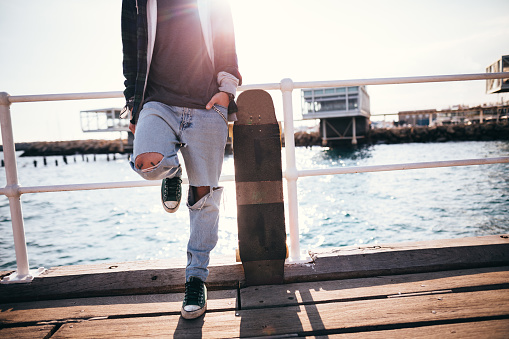 Young fashionable hipster skateboarder with longboard standing on pier and waiting by the sea