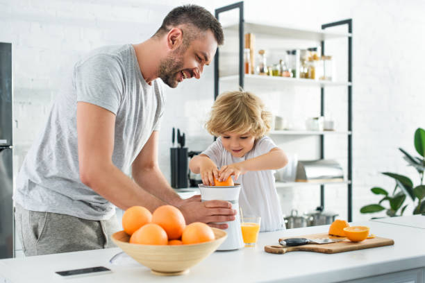 微笑的父親和小兒子做新鮮橙汁由擠壓在廚房桌上 - 榨汁機 個照片及圖片檔